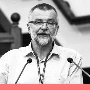 Black and white portrait of a man. He is wearing glasses, has a short beard and short-cropped hair. The man speaks through microphones and wears a shirt with a card around his neck.