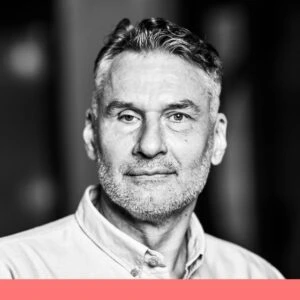 This image shows a man with short grey hair and a beard. He is wearing a light-coloured shirt and is looking directly into the camera with a neutral facial expression. The image is in black and white